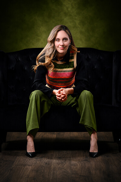 Allexis Tuccio, MCC, ACTC, EMCC, seated on a black sofa against a dark green studio background, looking directly at the camera.